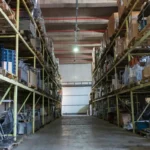 Lorong lebar gudang industri dengan rak logam bertingkat yang tersusun rapi, menggambarkan contoh layout gudang yang efisien